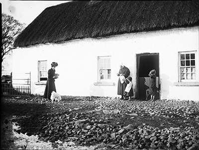 Edith Dillon carrying a box camera, approaching woman and 2 children standing outside thatched cottage