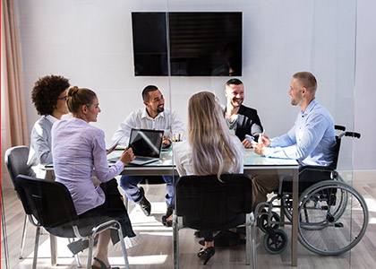Six business people sit in a meeting room, a mix of men and women of different backgrounds. One person uses a wheelchair. A laptop is on the table.
