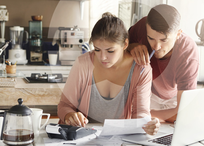 A couple review their household finances. The woman sits a table with the kitchen in the background while the man stands close to her. A laptop, calculator and many documents are in front of them.